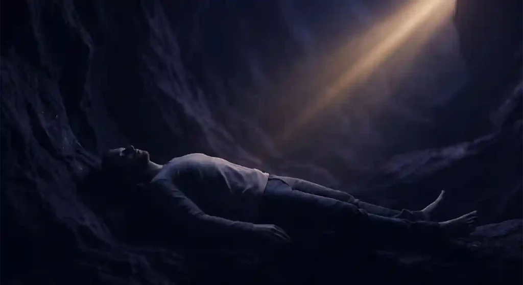 Imagen cinematográfica de un hombre acostado en el suelo de una cueva oscura, con un solo rayo de luz cálida iluminando su rostro y torso desde arriba.
